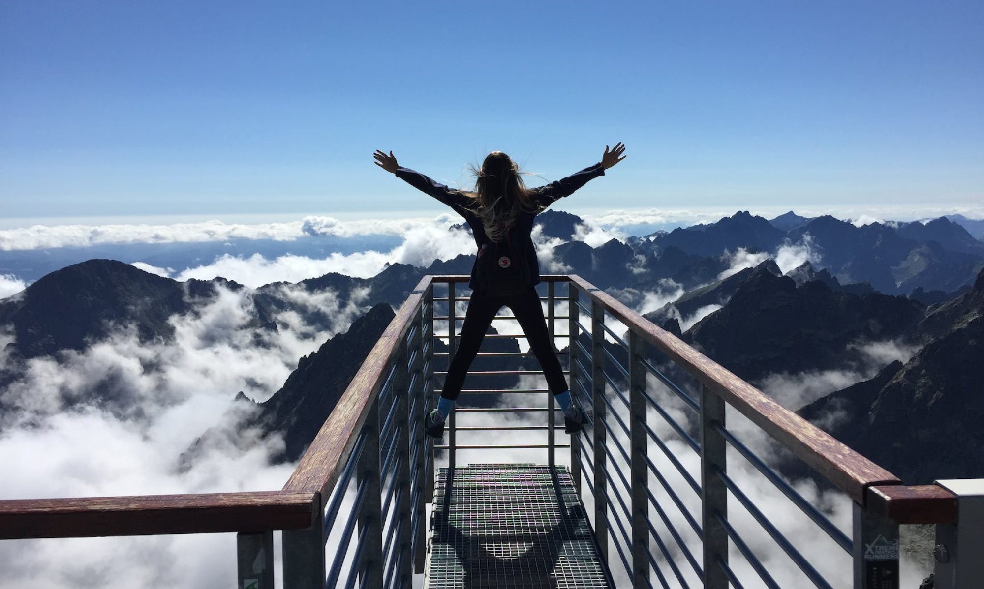 person standing on hand rails with arms wide open facing the mountains and clouds